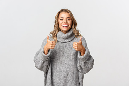 穿着灰色毛衣的快乐满意女性形象 展示拇指和微笑 推荐一些东西 比如并批准产品 站在白色背景上图片