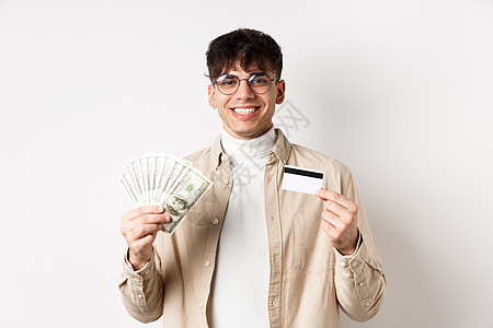 快乐的人拿着钱和塑料信用卡站着 年轻人拿着现金 满意地微笑 站在白色背景上账单黑发眼镜发型男人信用学生成人购物卡片图片