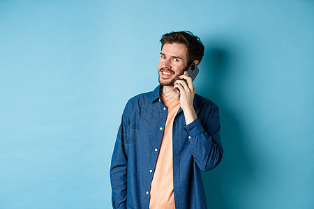 现代笑笑的家伙在手机上说话 看着快乐的相机 站在蓝背景上成人广告男人商业工具技术电话促销工作室成功背景图片