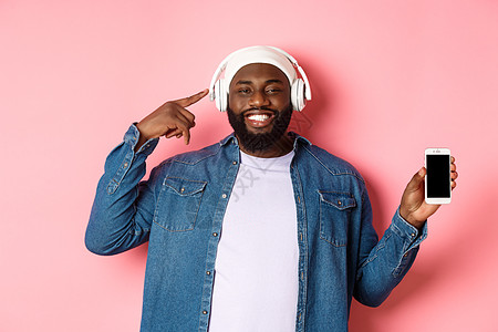快乐的黑人听音乐 指着耳机微笑 展示手机屏幕应用程序或播放列表 站在粉红色背景上网络学生促销帽子互联网购物男人胡子广告衬衫图片