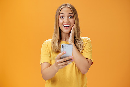 令人惊讶和印象深刻的女性对智能手机中令人敬畏的应用程序做出反应的肖像 惊讶和喜悦地对着相机手持手机在橙色墙壁上大笑手势震惊学生情图片