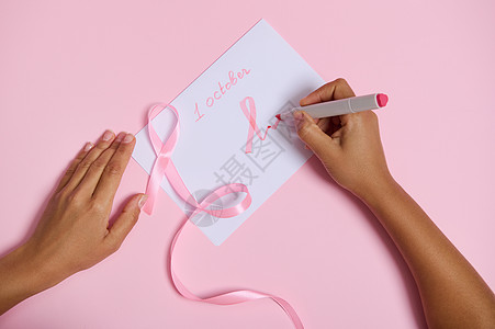 一位女士手握毡尖笔的顶部视图写下了 10 月 1 日 并在纸上画了一个粉红色的乳腺癌宣传月标志 一条粉红色的丝带 末端无尽 躺在图片