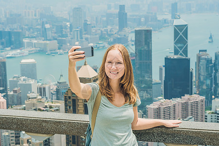 香港太平山女人用智能手机拍自拍杆照片 欣赏维多利亚港的景色 香港凌霄阁顶部的观景台 散焦背景 旅游亚洲概念女士港口观光天际城市景图片