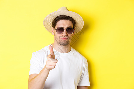 旅游和度假的概念 穿着夏日帽子和太阳镜的帅哥特写镜头指着相机 站在黄色背景上男性广告情绪微笑商业工作室假期游客手势旅行背景图片