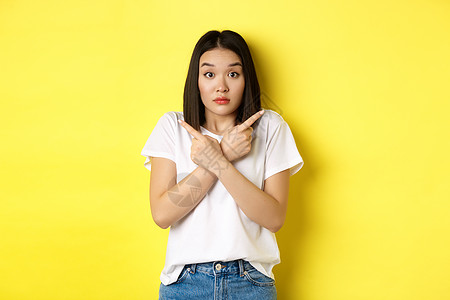 麻木不仁的亚洲女孩需要帮助选择 指指手指侧面和看不清 站在黄色背景上站立成人快乐理发黑发交易商业情感工作室广告青少年图片