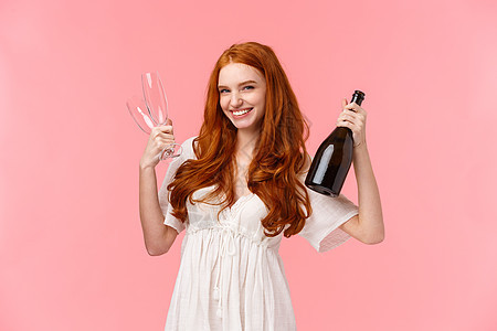 时髦漂亮的红头发女人和女朋友一起庆祝 一起玩乐 拿着一瓶香槟和两杯酒 邀请喝酒 面带笑容 开心 站着粉红色的背景图片