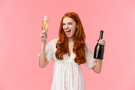 兴奋而无忧无虑的醉酒红发白人女友玩得开心 在很棒的派对上狂野而自由 举起玻璃杯和瓶装香槟 微笑时髦 站着粉红色背景图片