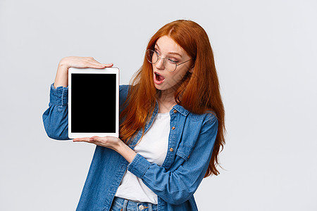 穿着牛仔衬衫的惊讶而好奇的可爱红发女性 看着惊讶的数字平板电脑屏幕 说哇 看看一些很棒的东西 站立的白色背景图片