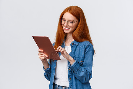 戴着眼镜的红头发 在数字平板电脑上看风趣和欢乐 笑高兴 对结果满意 站立白背景的Waist -up 肖像免费照顾的欧洲妇女图片