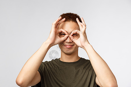 人 不同的表达方式和生活方式的概念 俏皮帅气的亚洲男人的画像 带着愉快的微笑透过手指眼镜看 做面具 玩得开心 灰色背景促销冒充互图片