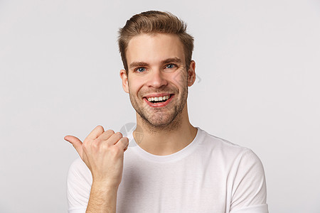 特写工作室肖像金发英俊和快乐 微笑的男人向左指着拇指询问朋友的意见 讨论公司产品 笑着开朗 站在白色背景商务男性技术企业家情绪胡图片