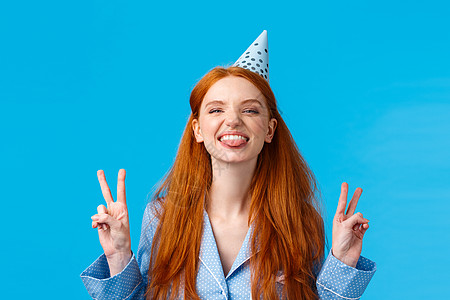 终于十八岁了开朗热情 无忧无虑的红头发女人 长发 雀斑 穿着睡衣 b-day 帽 舌头快乐 做出和平手势 站在蓝色背景图片