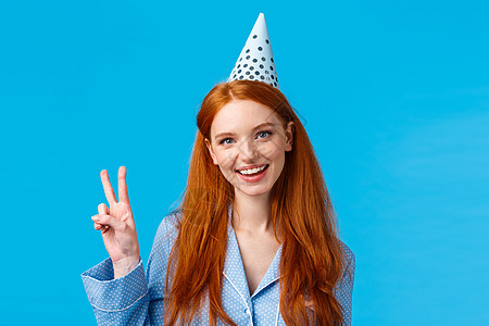 幸运而热情的女性漂亮红发女人 长着卷发 穿着睡衣 戴着显示和平标志的生日帽 快乐地微笑着 庆祝 b 日 蓝色背景图片