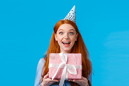 兴奋的漂亮女性 迷人的红发女人 蓝眼睛快乐的表情 收到可爱的粉红色礼物 礼物包裹在盒子里 笑得开心 用生日帽庆祝生日图片