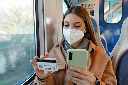 戴防护面罩的年轻女性乘坐火车旅行时用智能手机信用卡支付在线购物的画像图片