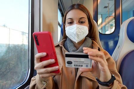 携带FFFP2 KN95保护面罩的妇女肖像 由火车在网上购物时用智能电话用信用卡支付费用图片