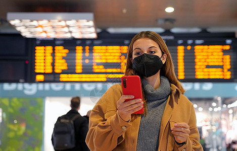 旅行妇女身着KN95 FFP2在下游站戴面罩 离开时有抵达时间的年轻女性在火车站内用手机检查信息 (单位 千斤顶)图片