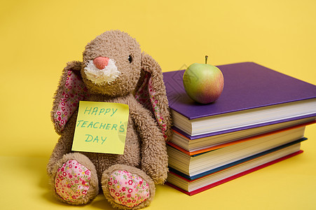 一只毛绒玩具兔子 在黄色便条纸上写着“教师节快乐” 靠在一堆彩色书籍上 黄色背景 有文字空间图片
