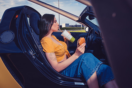 穿着蓝色牛仔裤和橙色顶部的女士喝纸杯饮料 坐在黄色轿车上时拿着汉堡包 快餐 装饰图片