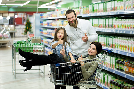 美丽的年轻父母和可爱的小女儿在超市中选择食物时微笑着 父亲推了购物车 而女孩们却坐在那里等他(她)健康高清图片素材