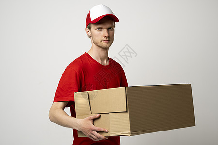 身穿红色制服的送货员和拿着纸箱的帽子 装有邮政包裹 准备用白色工作室背景向客户发送 (笑声)图片