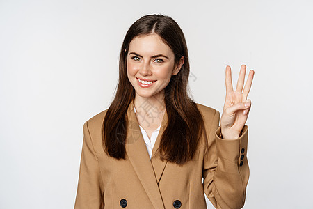 公司女性的肖像 女售货员展示了三号手指和笑容 穿着西装站在白色背景上图片