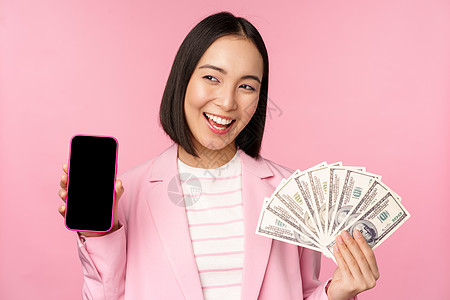 成功的年轻亚洲女商务人士展示金钱 现金和智能手机屏幕 微笑着高兴 粉红色背景售货员办公室信用贷款银行人士细胞购物学生工作室图片