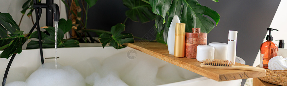 用于美容和身体护理产品的木架 位于现代白色泡泡浴缸上方的木架上 用绿色热带植物龟背竹装饰的舒适时尚的浴室洗发水瓶子风格凝胶房间化图片