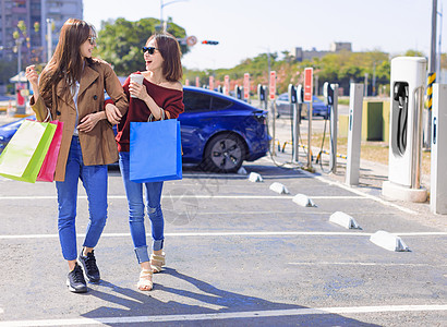 快乐的年轻女人站在电动车附近的城市停车场 从小城市车站给汽车电池充电 拿着购物袋 一起走购物运输插头技术活力力量驾驶充电器发动机图片