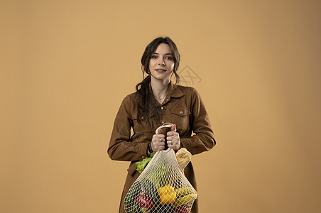 快乐微笑的年轻女子的画像 穿着棕色连衣裙 拿着可重复使用的网袋 里面装着杂货 背景是橙色的 可持续性 生态生活和人们的观念网兜食图片