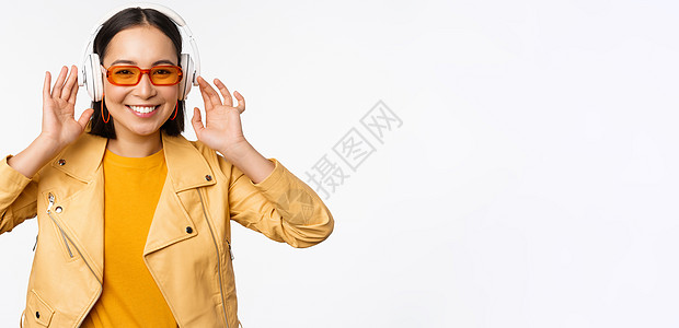 美丽的亚洲女孩 欢笑快乐 用耳机听音乐 站在白色背景上站立情绪女士成人快乐办公室太阳镜企业家女朋友工作购物背景图片