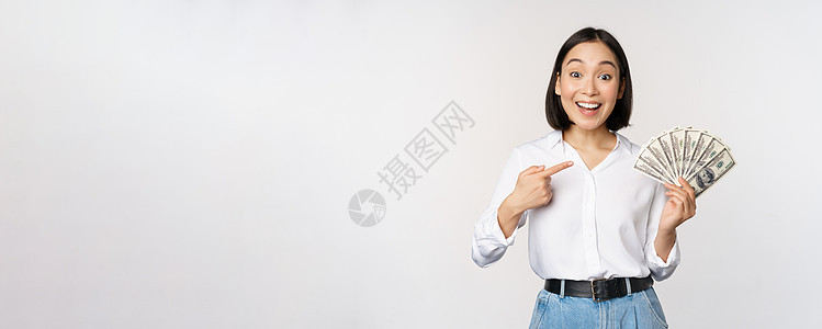 年轻亚洲女人的肖像 指着她的钱币 显示现金 站在白种背景上银行工作室商业快乐冒充人士技术女孩女士信用图片
