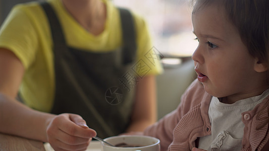 特写妈妈用白色喂养小婴儿喂养高脚椅 孩子试图自己吃东西 快乐的孩子脸上沾满食物 小女孩用勺子吃粥 高品质的厨房早餐喜悦营养手工母图片