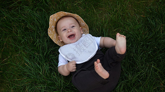 夏日阳光明媚的夏日 戴着夏日巴拿马帽的小快乐新生儿赤脚躺在草地上 在家庭童年自然之外的花园里 婴儿小孩蹒跚学步的男孩微笑着看着相图片