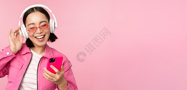 时尚的亚洲女孩用智能手机跳舞 在手机应用程序上戴着耳机听音乐 笑着笑着 在粉红色背景下摆姿势音乐办公室促销黑发情感广告互联网成人背景图片