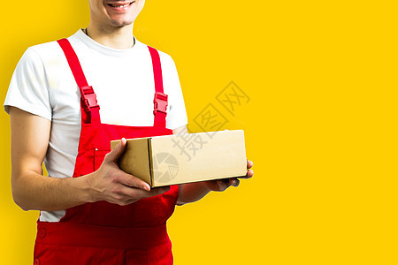 一个带着红帽子的快乐年轻送货员 与黄色背景隔绝的包裹邮箱一起站立图片