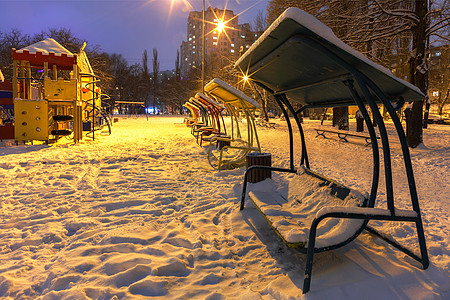 冬季城市夜晚公园的木凳和游乐场在蓝色黄昏的背景下被雪覆盖着 (笑声)图片