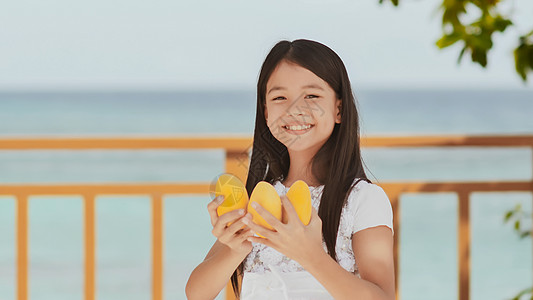 一个穿着白色裙子的迷人的菲利平女学生 长发正装着芒果 在她手中 阳光 蓝海 童年 娱乐 笑声微笑牛奶食物享受橙子孩子小吃营养果汁图片