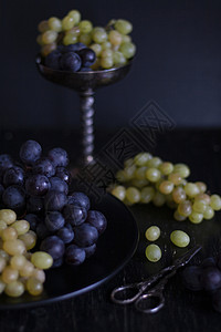 一串深蓝色和绿色的葡萄躺在一个黑色的盘子里 立在一个银色的碗里 旁边是葡萄剪刀 在黑色背景上的组成古董浆果水果明信片花瓶食物餐具图片