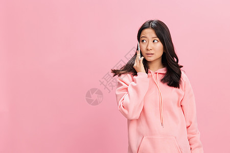 身穿粉色连帽衫运动衫 困惑 不快乐 可爱的亚洲学生年轻女士从粉红色工作室背景中孤立的电话中得知悲伤的消息 好报价 小工具成瘾社交背景图片