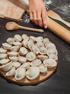 女人做 pelmeni 或饺子-俄罗斯传统菜肴 由面团和肉制成 有面粉和木器物的黑色厨房桌子图片