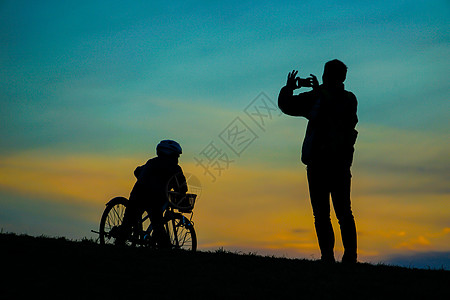 父母和子女在日落山上站立的轮椅自行车爬坡头盔公园字符父亲太阳日落美景草原图片