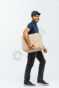 送货概念  穿着蓝布的快乐非洲裔美国送货员的画像 他走着向顾客发送包裹 隔绝在灰色演播室背景 复制空间邮件商业服务邮递员盒子运输图片