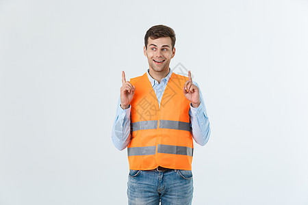 身穿橙色背心的灰墙上的英俊年轻专业工程师男子惊讶地看 用手指指着 笑声工人安全安全帽男人快乐承包商建设者成功工作惊喜图片