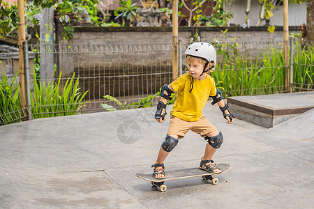 滑板男孩戴头盔和膝盖垫的体育男孩学会在滑板公园与滑板一起玩滑板孩子运动公园乐趣游戏男生街道平衡驾驶城市生活背景