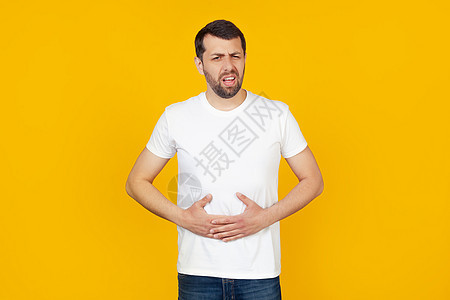 一个留着胡子的年轻男子穿着白色 T 恤 由于消化不良 手放在肚子上 感觉不舒服 痛苦的概念 站在孤立的黄色背景上中毒悲伤伤害男性图片