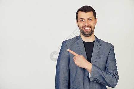 留着胡子的年轻商务人士穿着夹克 开朗 脸上带着微笑 用手和手指指着一边 表情快乐而自然 灰色背景下的男人肖像相机广告手势白色成人图片