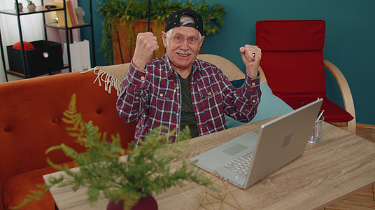 祖父在笔记本电脑上玩电脑电子游戏 赢得 庆祝 欢笑和快乐的微笑图片