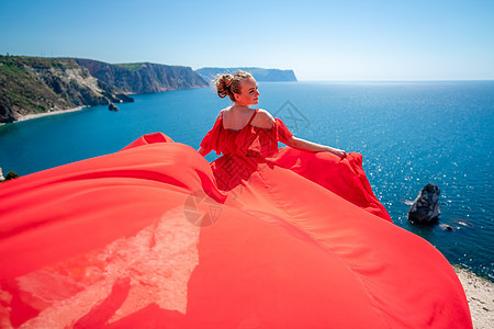 金发碧眼的长发在阳光明媚的海滨穿着红色飘逸的连衣裙 后视 丝绸面料在风中飘扬 在海边的蓝天和群山的映衬下衣服女孩太阳飞行幸福旅行图片
