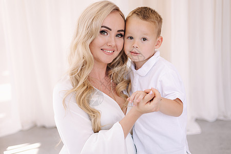 男孩拥抱他的妈妈 穿着优雅的白色裙子的女士在摄影棚里给摄影师摆上姿势 白发背景 (笑声)图片